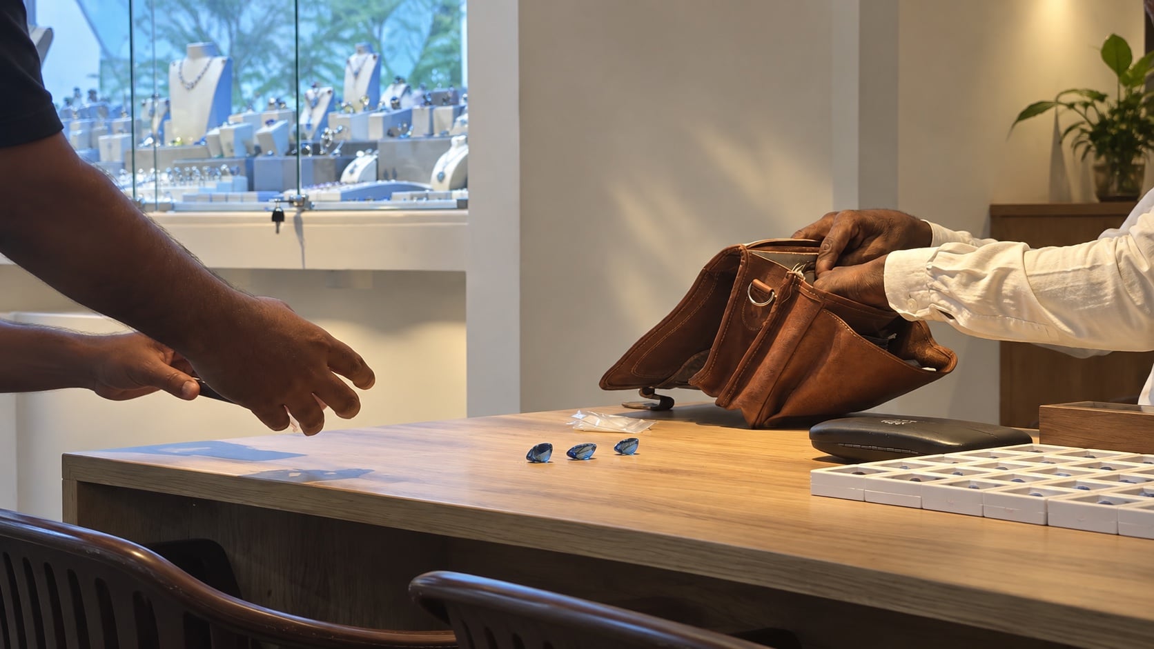 Two pairs of hands over a wooden counter: three blue sapphires laid out, an open leather satchel and a stone tray
