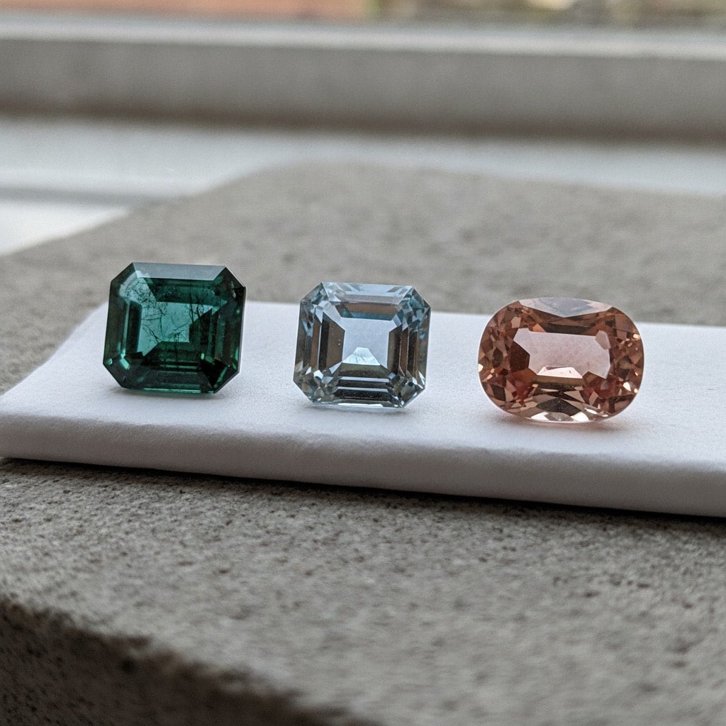 An emerald-cut green emerald, an emerald-cut pale blue aquamarine and an oval peach morganite side by side on gem paper, three colours of one mineral species
