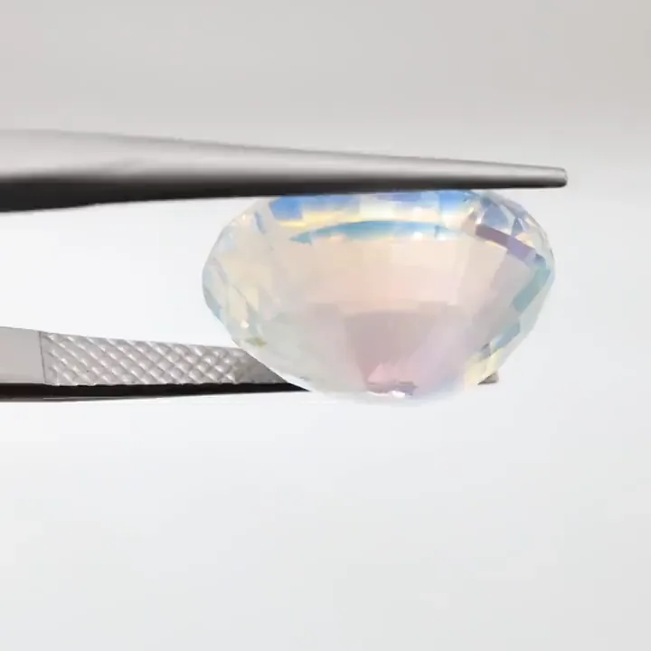 Une pierre de lune arc-en-ciel non sertie, taille ovale, 11,11 ct, tenue à la pince sur fond gris, un reflet bleu sous ses facettes