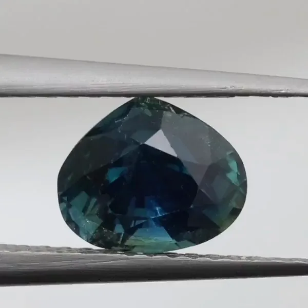 A pear-cut sapphire held in tweezers and turned: deep blue seen through the top, a lighter blue-green seen from the side