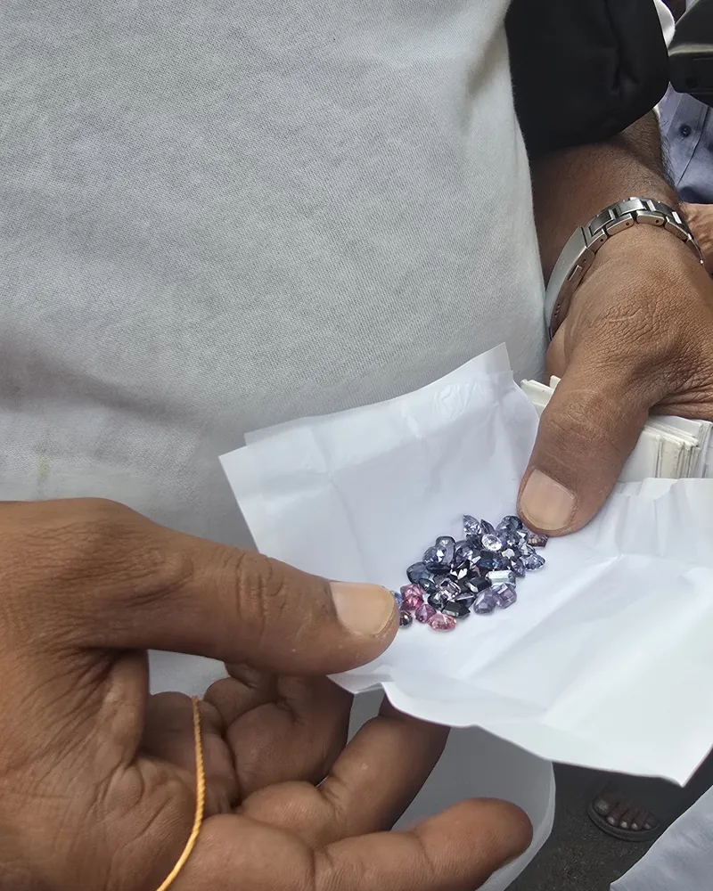 Hands holding an open paper fold of small blue, violet and pink cut stones in a street market