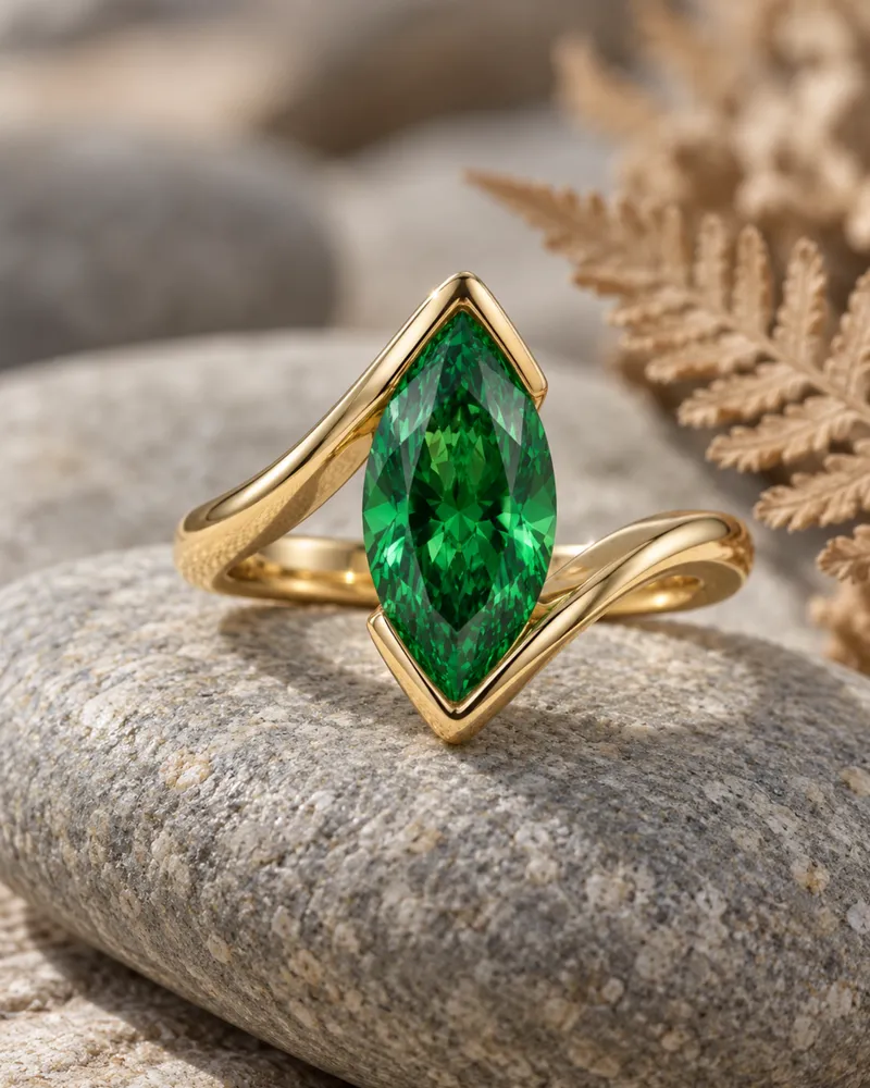 Illustration d’une bague croisée en or avec un grenat vert marquise tenu par le métal à ses deux pointes, posée sur un galet lisse.