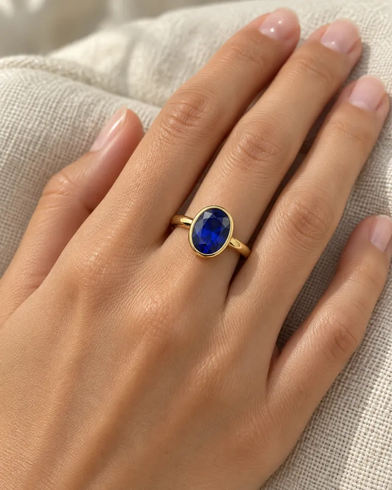 Illustration d’une bague en or en serti clos avec un saphir bleu roi ovale, portée sur une main posée sur un coussin de lin.