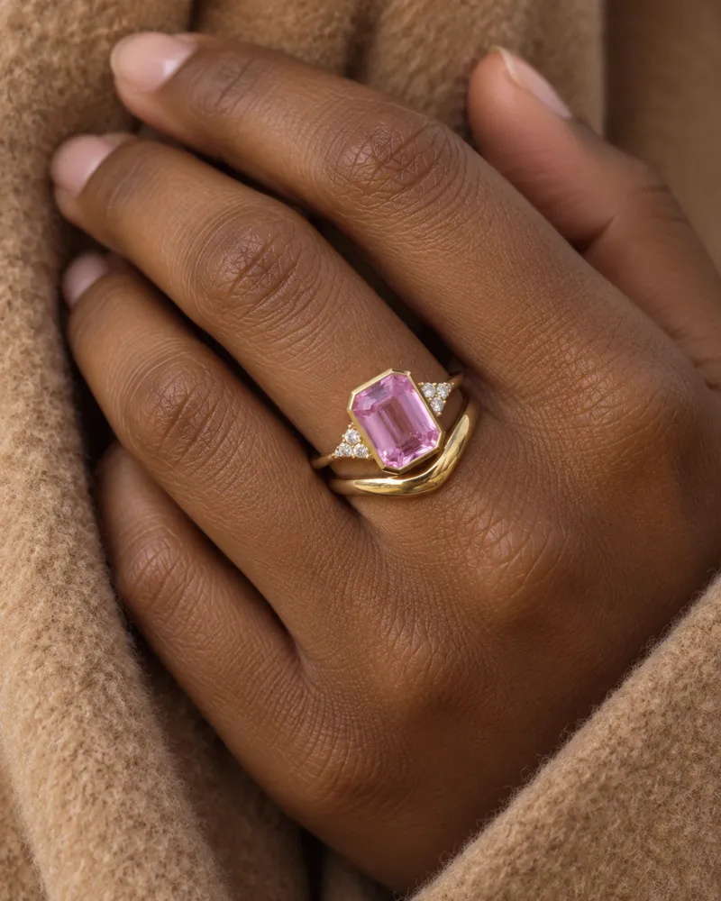 Illustration d’une bague en or en serti clos avec un saphir rose taille émeraude et de petites pierres latérales, portée avec une alliance en or galbée, la main contre un manteau de laine camel.