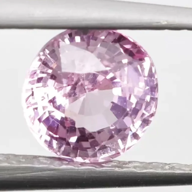 Loose sapphire listed as pastel baby pink, 1.05 ct, round cut, held in tweezers on a light grey ground