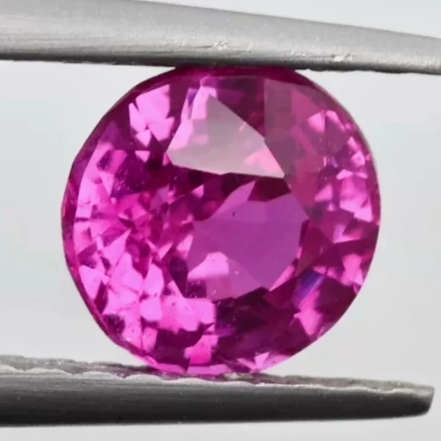 Loose sapphire listed as purplish pink, 0.97 ct, round cut, held in tweezers on a light grey ground