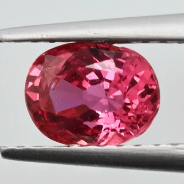 Loose sapphire listed as reddish pink, 0.95 ct, oval cut, held in tweezers on a light grey ground